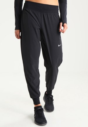 Person trägt lockere schwarze Nike-Jogginghosen und schwarze Sportschuhe und steht vor hellem Hintergrund.