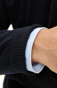 Granatowy sweter dzianinowy z prążkowanymi mankietami nałożony na jasnoniebieską koszulę, charakteryzujący się gładką fakturą i wyraźnym kołnierzykiem z subtelnym wzorem kratki.