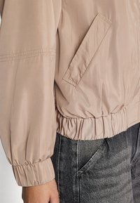 Veste bomber rose clair avec des poignets et une ceinture élastiques, dotée d'une poche latérale et d'une texture lisse et brillante. Associée à un jean en denim noir.