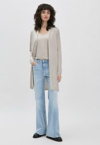 Beige lange cardigan met open voorkant, over een lichtgrijs topje. Gepaard met lichtblauwe flared jeans en zwarte schoenen. Zachte textuur.