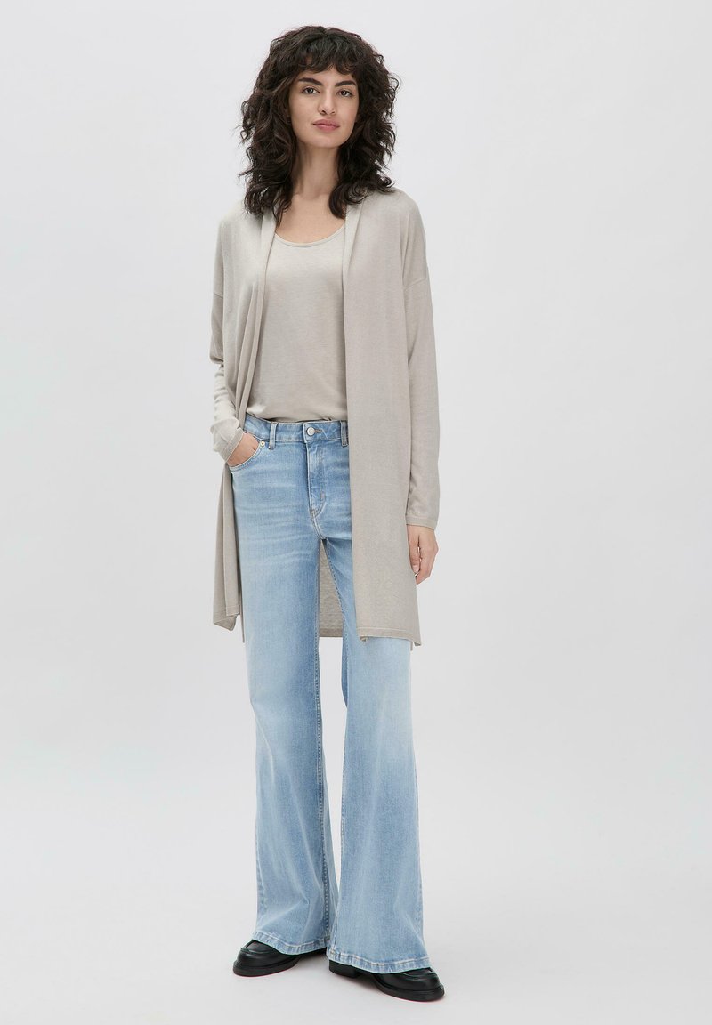 Beige lange cardigan met open voorkant, over een lichtgrijs topje. Gepaard met lichtblauwe flared jeans en zwarte schoenen. Zachte textuur.