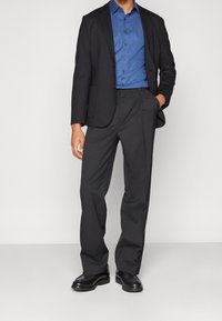 Giacca nera sopra una camicia button-down blu, abbinata a pantaloni scuri e scarpe nere. Tessuto morbido, vestibilità su misura, design classico.
