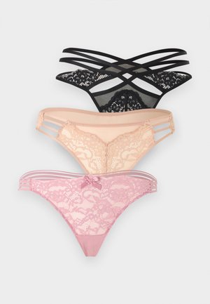 Femme aux longs cheveux foncés portant un soutien-gorge en dentelle beige et une culotte en dentelle assortie, debout, les mains derrière le dos, devant un fond uni.