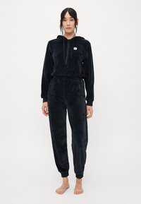 Tuta in velluto nero con felpa con zip e cintura elasticizzata. Presenta polsini a coste e dettaglio del logo frontale. Texture morbida in tutto il capo.