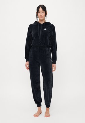 Zwarte fluwelen trainingspak met een hoodie met rits en een elastische tailleband. Bevat ribgebreide manchetten en een logo-detail aan de voorkant. Zacht aanvoelen door het hele pak.