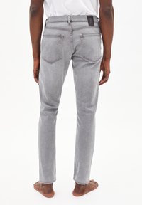 Jean en denim gris coupe slim, avec poches arrière et une étiquette noire à la taille. Le tissu a une texture lisse et des coutures minimales.