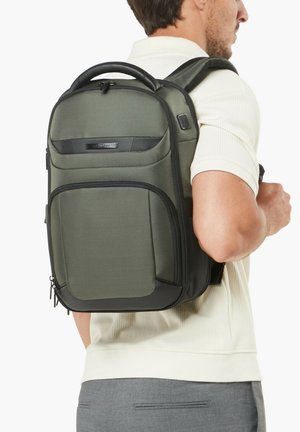 Mann trägt ein helles Hemd und graue Hose, hat einen dunkelgrünen und schwarzen Rucksack mit mehreren Reißverschlussfächern auf dem Rücken.