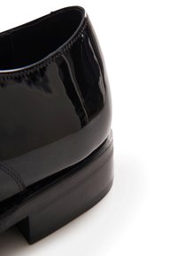 Scarpa in vernice nera con superficie liscia e lucida, punta squadrata e tacco impilato. Presenta cuciture sottili lungo il bordo superiore.