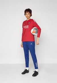 Röd långärmad tröja med Qatar Airways-logotyp, blå sportbyxor med texten "PARIS", svarta fotbollsdojor, håller en svartvit mönstrad fotboll.