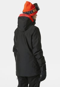 Černá voděodolná bunda s červeným akcentem na kapuci, s volným střihem, hladkou texturou a nastavitelnými manžetami. Je na sobě oranžová pletená čepice.