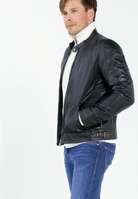 Zwarte leren jas met een gladde textuur, ritsclosure en drukknoopcollar. Draagt over een witte trui en gecombineerd met een blauwe spijkerbroek.