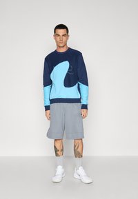 Sweatshirt azul-marinho e azul claro com design abstracto, combinado com calções cinzentos. O modelo usa ténis brancos e tem tatuagens visíveis nas pernas.