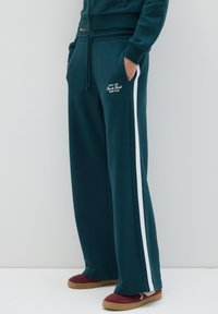 Persona que lleva pantalones de chándal de un verde azulado oscuro con franjas laterales blancas y un logo, con las manos en los bolsillos, acompañados de zapatos de slip-on color marrón burdeos con suelas beige.