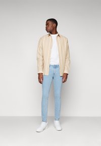 Camisa de botones de rayas beige sobre una camiseta blanca, combinada con jeans skinny azul claro y zapatillas blancas. Fondo neutro.