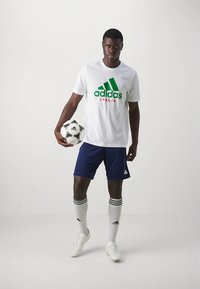 Camiseta blanca de Adidas con logo verde y rojo, pantalones cortos azules, calcetines blancos con rayas negras y zapatillas blancas, sosteniendo un balón de fútbol negro y blanco.