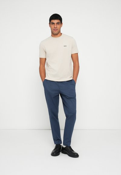 T-shirt beige avec logo noir, associé à un pantalon tailleur bleu marine et des chaussures à lacets noires. Tissu lisse, design simple, coupe décontractée.