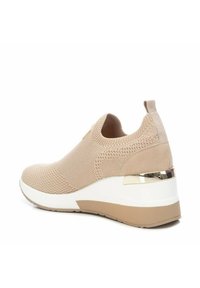 Zapatilla de punto beige con parte superior texturizada, suela blanca acolchada y acento metálico en el talón. Con una pestaña para facilitar el uso.