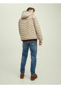 Beige péřová bunda s vzorem ve tvaru šipek, kapucí a elastickými manžetami, nošená s modrými džíny a hnědými botami, pohled zezadu.