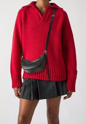 Personne portant un pull en maille rouge, une jupe plissée grise, un sac bandoulière noir et un collier en argent sur un fond uni.