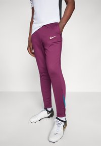 Nike sportnadrág sötétlila színben, slim fit fazonnal, nedvszívó anyaggal, oldalsó zsebekkel, és türkiz színű díszítéssel a szegély közelében.