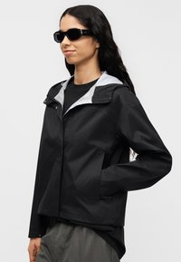 Μαύρο αδιάβροχο μπουφάν με γκρι επένδυση κουκούλας, πλαϊνές τσέπες και χαλαρή εφαρμογή. Επιπλέον, μαύρα γυαλιά ηλίου συμπληρώνουν το σύνολο.