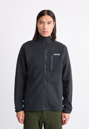 Black fleece zip-up jacket with a high collar and a chest pocket. Features two side pockets, textured fabric, and contrasting material details.