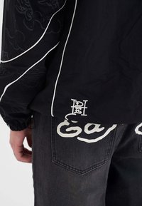Giacca nera con piping bianco e motivi sottili, abbinata a jeans scuri con grandi lettere bianche sul retro.