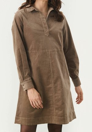 Femme portant une robe en velours côtelé marron à manches longues, avec un col et des boutons aux poignets, debout devant un fond uni.