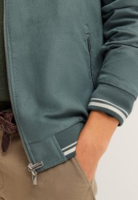 Hellblaugrüne Reißverschlussjacke mit perforierter Textur, gestreiften Bündchen und Details an den Seitentaschen. Kombiniert mit beigen Hosen.