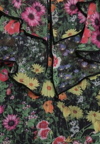 Tissu à motif floral avec des fleurs vibrantes rose, jaune et violettes sur un fond vert ; présente des bords noirs froncés et une texture transparente.