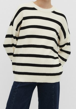 Personne portant un pull en maille crème avec de larges rayures horizontales noires et un jean bleu foncé, debout les mains derrière le dos.