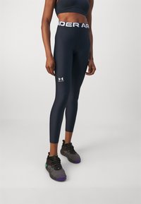 Granatowe legginsy z białym paskiem i logo "Under Armour". Odpowiednie do aktywności fizycznej, gładka tekstura, dopasowany krój. Czarne buty sportowe.