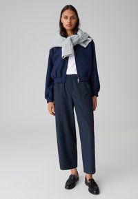 Veste de bombardier marine sur une chemise blanche, associée à un pantalon large marine. Une écharpe gris clair drape les épaules. Mocassins noirs.