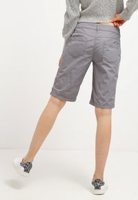 Pantalones cortos de algodón gris con un corte relajado y largo hasta la rodilla, dos bolsillos traseros y detalles de costura, combinados con zapatillas blancas con manchas grises.