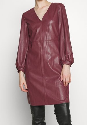 Frau trägt ein langärmeliges, burgunderfarbenes Kunstlederkleid mit V-Ausschnitt und schwarze Overknee-Lederstiefel vor schlichtem Hintergrund.