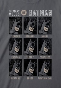 Grijze t-shirt met negen illustraties van een gemaskerde figuur met verschillende gezichtsuitdrukkingen: serieus, ontspannen, boos, opgewonden, slaperig, verward, neutraal, verveeld en vechtend tegen het kwaad.