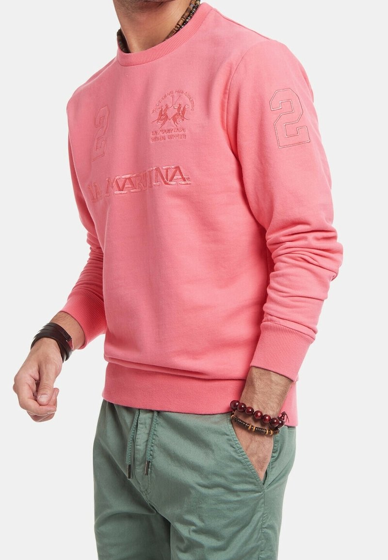 Růžová mikina s kulatým výstřihem, zdobená texturovanými detaily loga a velkým číslem "2" na rukávu. Kombinováno se zelenými kalhotami.