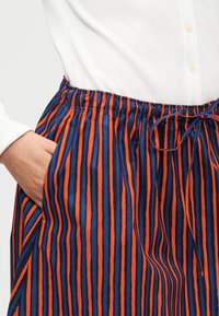 Μέση και χέρι στην τσέπη μιας ριγωτής πορτοκαλί και μπλε φούστας με ελαστική μέση, συνδυασμένη με λευκό ριμπ μακρυμάνικο πουκάμισο με κουμπιά.