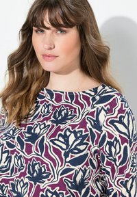 Mladá žena se světle hnědými vlnitými vlasy, oblečená v topu s květinovým vzorem v odstínech fialové, námořnické modři a bílé, s knoflíky poblíž výstřihu.