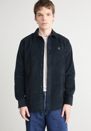 Camisa de pana negra de botones con cuello extendido, logo bordado en el pecho y mangas largas, combinada con jeans azules.