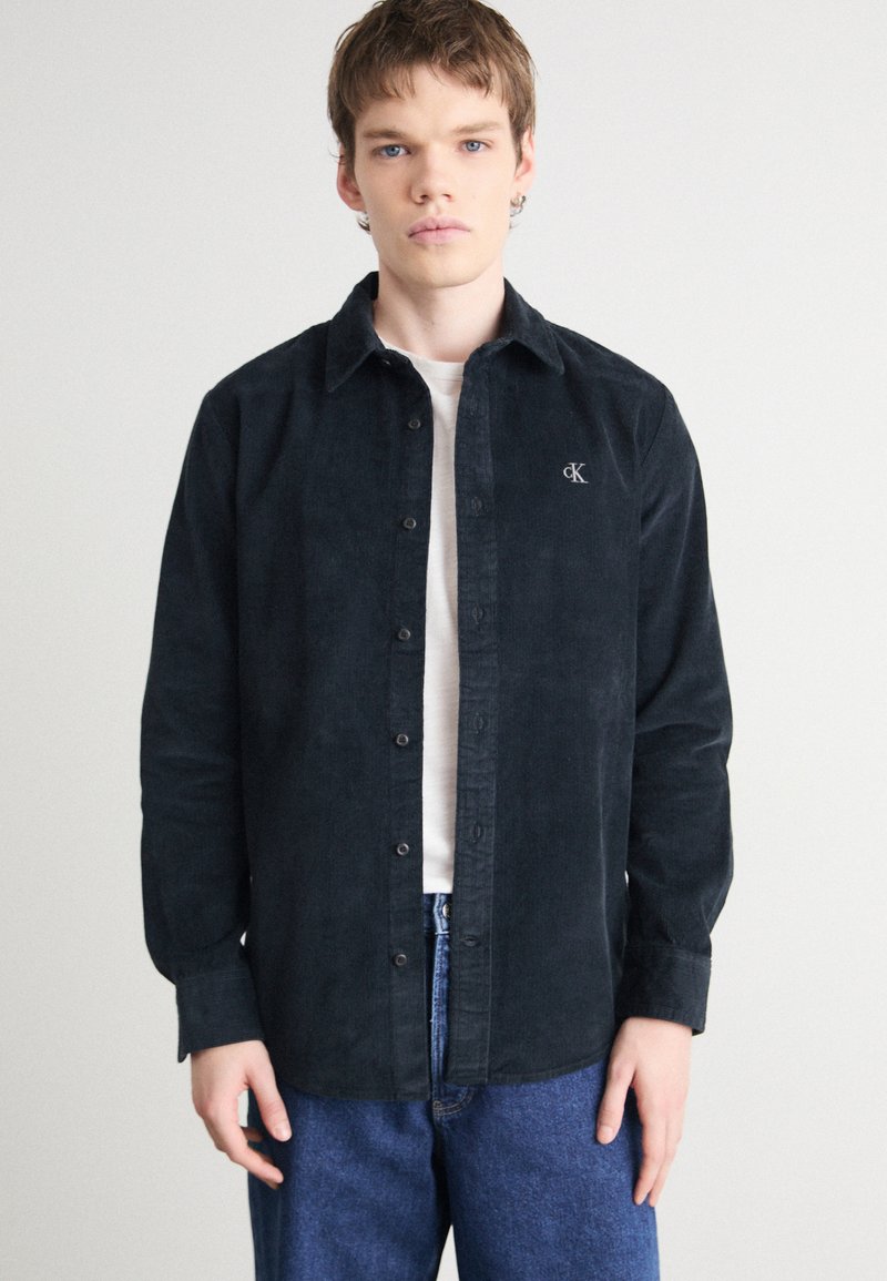 Camicia button-up in corduroy nera con colletto a punta, logo ricamato sul petto e maniche lunghe, abbinata a jeans blu.