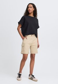 Svart, kortermet, løs passform topp paret med beige cargo shorts med sidelommer, rullede ermer og svarte og beige sneakers.