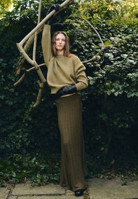 Maglione a coste verde oliva e gonna lunga abbinata. Tessuto testurizzato con sottili righe verticali. Guanti di pelle nera e calzature lisce.