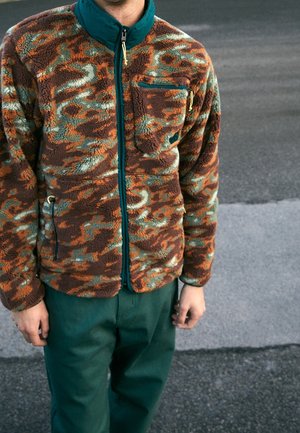 Personne portant une veste polaire camouflage marron, verte et orange avec un pantalon vert foncé, debout sur une surface pavée en extérieur.