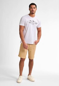 Bílé bavlněné tričko s grafikou auta a textem, doplněné o béžové šortky a světlé tenisky. Model stojí na světlém pozadí.