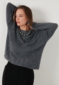Grau gestrickter Pullover mit lockerer Passform, der auffällige horizontale Löcher und einen runden Ausschnitt aufweist. Kombiniert mit einer Halskette aus metallischen Perlen.