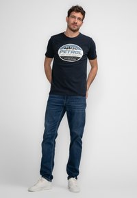 Marineblauw katoenen T-shirt met een witte bedrukte afbeelding van "PETROL" en berggraphics, gecombineerd met een blauwe spijkerbroek en witte sneakers.