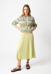 Spalvota raštuota bomber tipo striukė su žaliais, mėlynais ir oranžiniais akcentais ant šviesiai žalios plisuotos suknelės. Išvaizdą užbaigia rudos virviškos šlepetės.