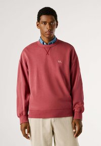 Sudadera roja con puños y dobladillo acanalados, que presenta un detalle de punto cruz en el cuello y un logo blanco "PJL" en el pecho.