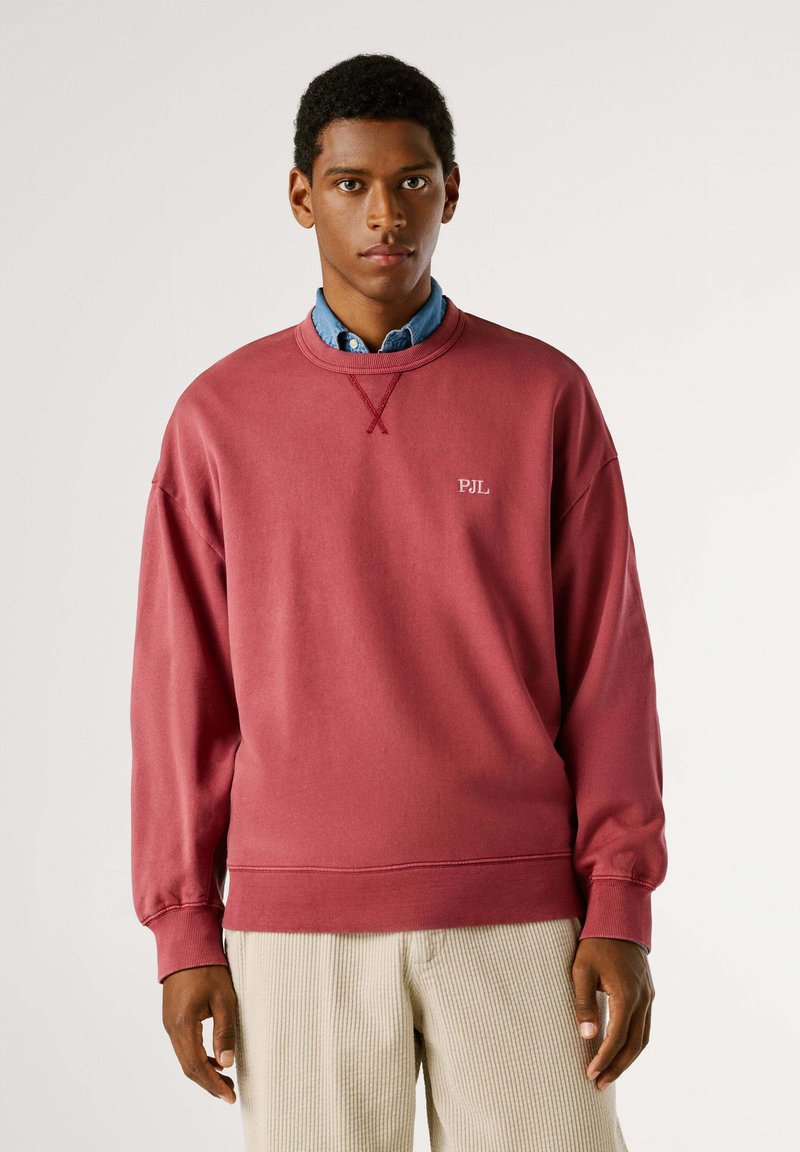 Sudadera roja con puños y dobladillo acanalados, que presenta un detalle de punto cruz en el cuello y un logo blanco "PJL" en el pecho.